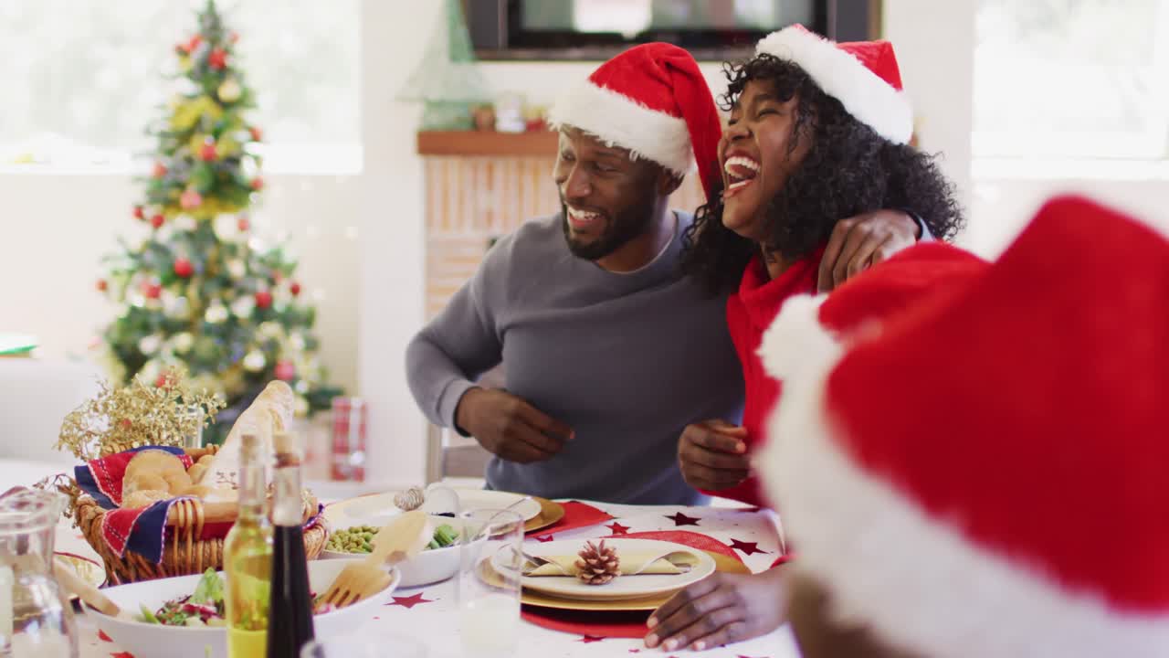 Coppia afroamericana con cappelli di Babbo Natale che sorride e brinda mentre è seduta a tavola a pranzare.