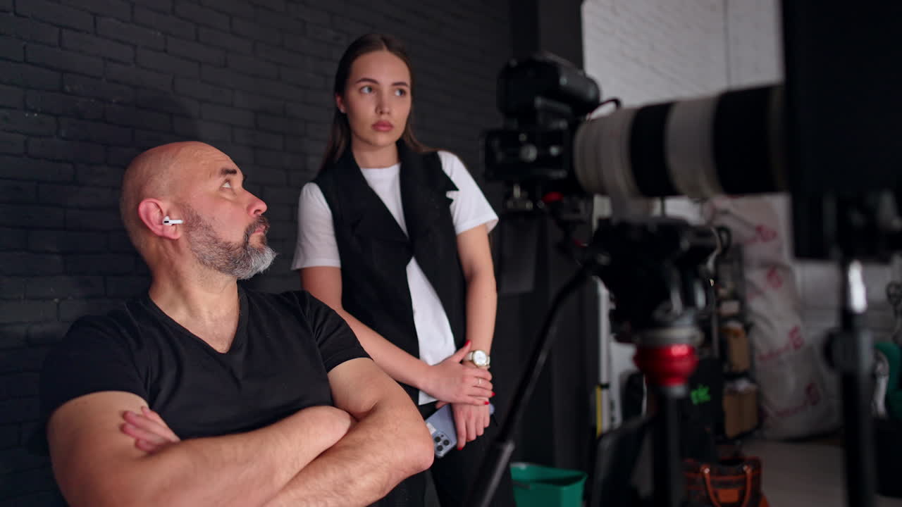 Bearded photographer and young lady are looking at camera display. Filming crew working in the studio.