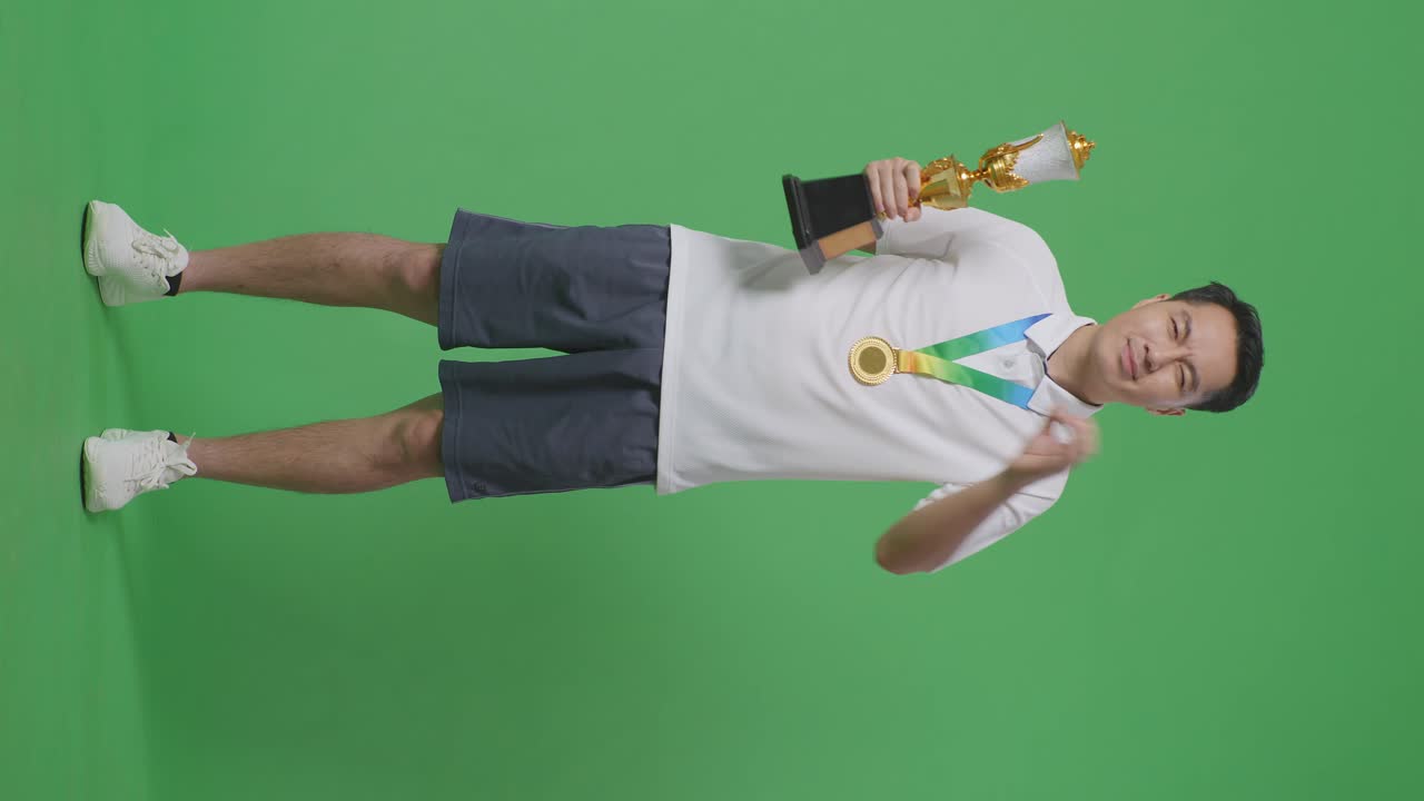 금메달과 트로피를 가진 아시아 남자의 몸이 가득 차 있습니다. 오케이, 손을 눈 위에 고 카메라에 미소 짓고 있습니다. 스튜디오의 녹색 스크린 배경에서 첫 번째 우승자로서.