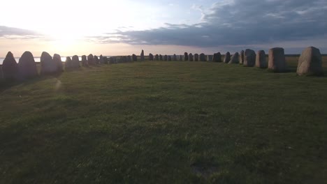 Through-Ancient-Oval-Formed-Stones-Ales-Stenar-By-The-Sunset-in-South-Sweden-Skåne-Österlen-Kåseberga,-Aerial-Very-Low,-Close-To-Ground-Forward