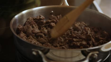 cooking brown meat pieces in silver pot on kitchen stove with wooden slatted spoon, close up static