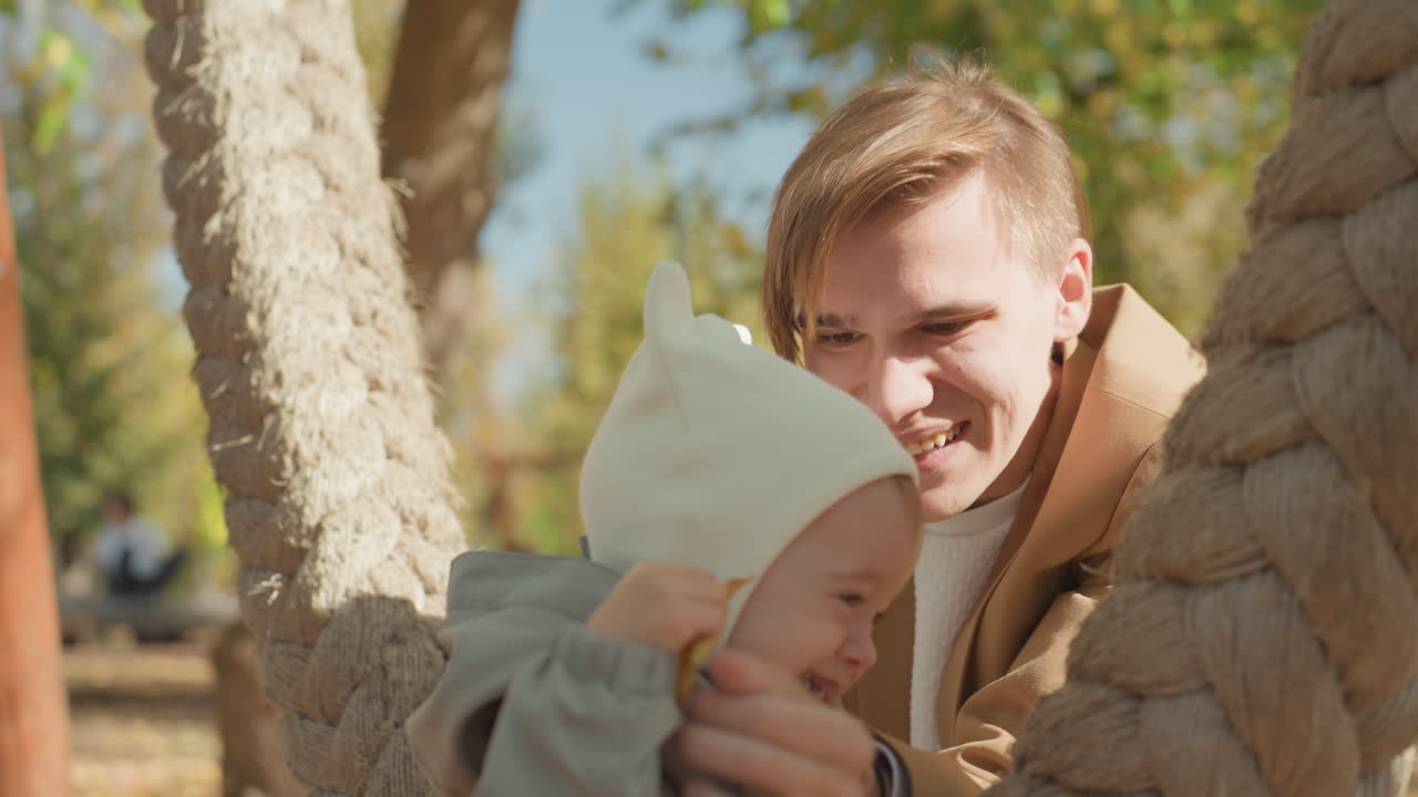 Herbstpark, Vater und Kleinkind spielen auf einer Seilschaukel; warmes Sonnenlicht filtert durch goldene Blätter; Vater hockt und ermutigt das Baby, sich auszustrecken; gemütliche Strickmütze, ehrliches Lächeln und Zärtlichkeit.
