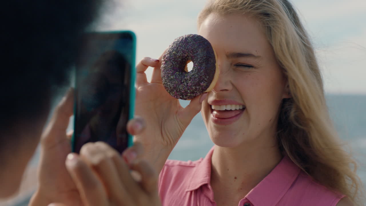 hermosa mujer posando con una rosquilla en la playa mejor amiga tomando fotos usando un teléfono inteligente compartiendo fin de semana en la playa en las redes sociales disfrutando de la diversión de verano 4k