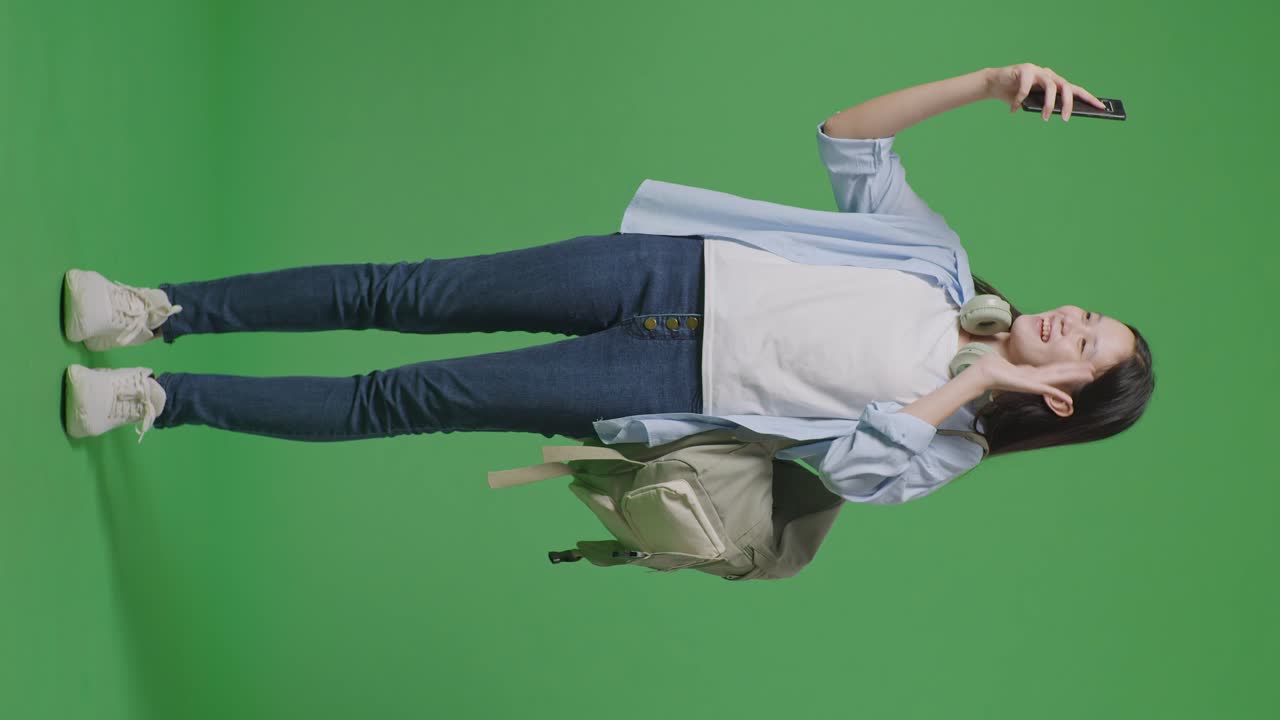 녹색 화면 배경 스튜디오에 서있는 동안 스마트폰에서 비디오 통화를하는 배과 몇 권의 책을 가진 아시아 십대 소녀 학생의 몸이 가득 차 있습니다.