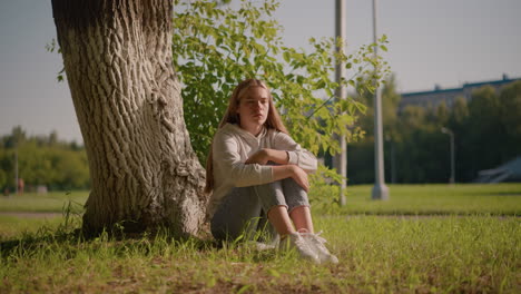 dama se sienta en la hierba con los ojos cerrados, descansando por el tronco del árbol en un momento pacífico, la luz del sol se refleja suavemente en ella, los brazos cruzados sobre las rodillas, el fondo incluye el estadio, los postes, y el corredor lejano