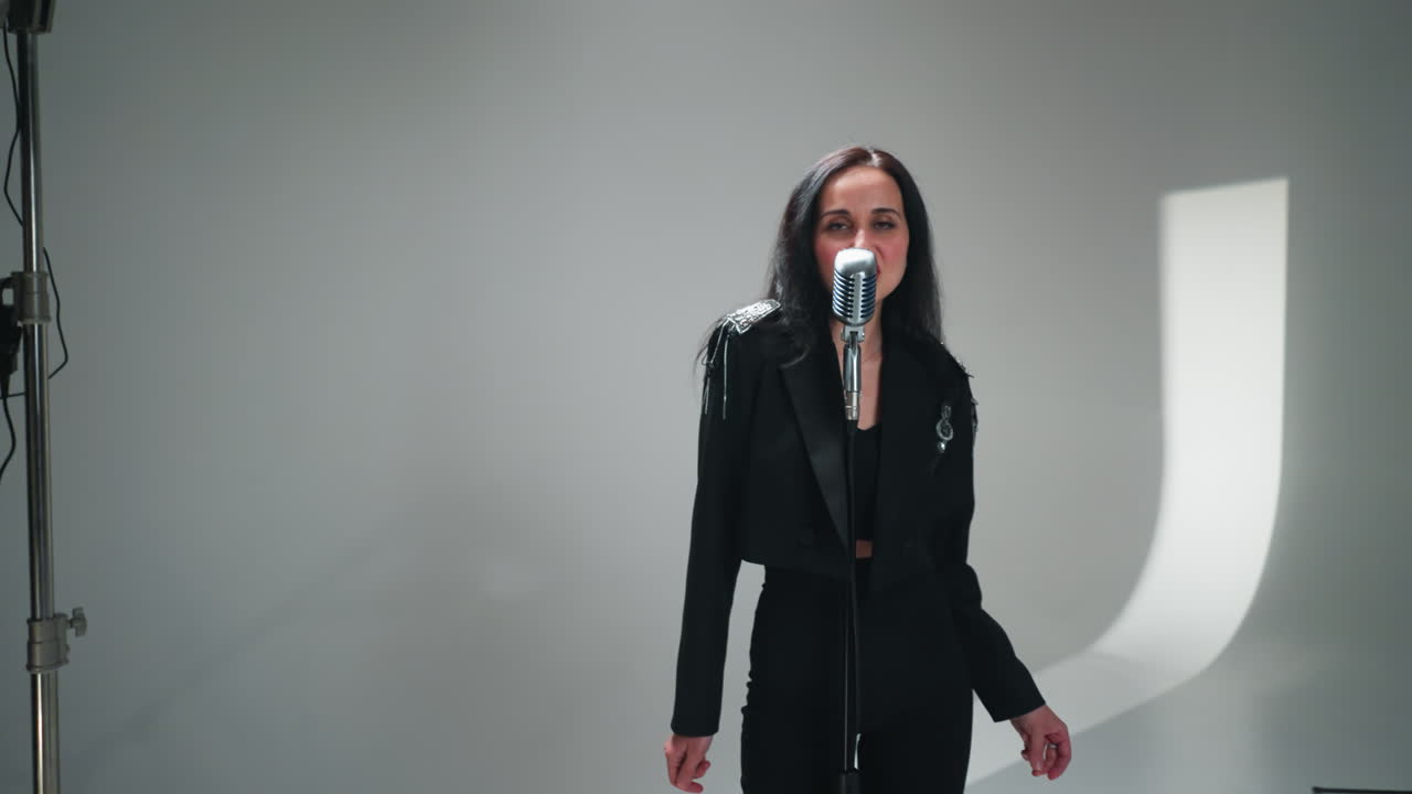 검은 옷을 입은 우아한 여성 보컬리스트가 카메라가 아지는 배경에서 키보디스트와 함께 빈티지 마이크에서 공연합니다.