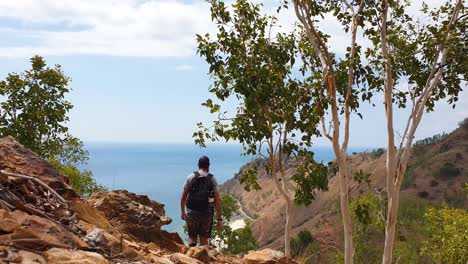 Hombre-Caminando-Por-El-Desierto-Al-Aire-Libre-En-Las-Colinas-Con-Terreno-Rocoso-Con-Vista-Al-Hermoso-Océano-Azul-En-Un-Remoto-Destino-De-Isla-Tropical,-Alejándose-De-La-Cámara-Hacia-La-Vista-Del-Mar