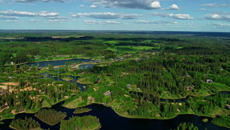 Drone-flying-above-the-picturesque-landscape-of-Amatciems,-Latvian-eco-village-carved-through-lake-and-lush-green-trees