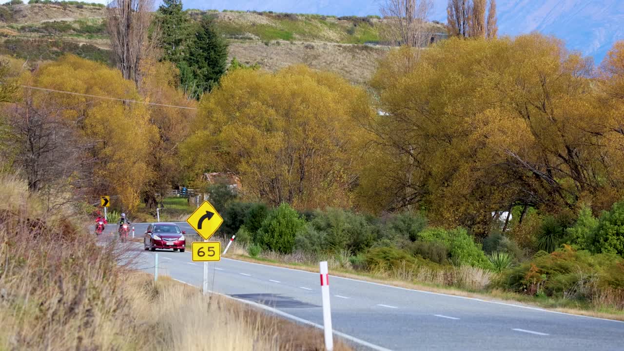 A car navigates a winding road surrounded by vibrant autumn foliage in Glenorchy, New Zealand, under clear daylight