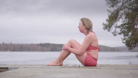 emotional and vulnerable woman in swimsuit sits on lake pontoon after ice bath