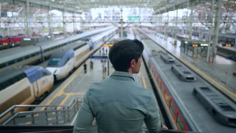 rear view of man moving down on escalator at seoul ktx train station overlooking many railway speed trains arriving and waiting for departure - high rise slow motion