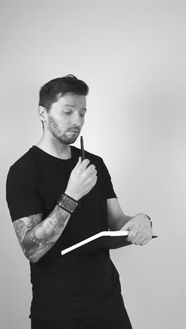 grayscale footage of a tattooed man thinking about something with putting a pen on his chin and looking up, then continues to write something down in the black journal that he is holding in his hands.