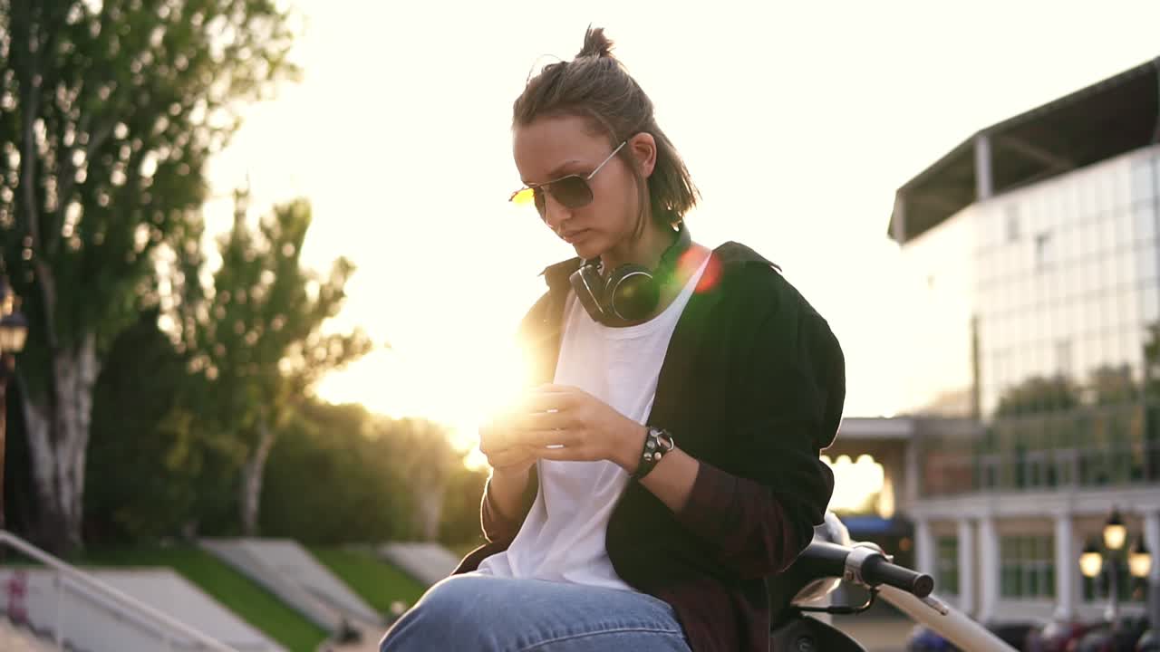 una chica moderna y a la moda se sienta en un ciclomotor y envía mensajes de texto en su teléfono móvil. lleva vaqueros, un jersey blanco y un suéter negro