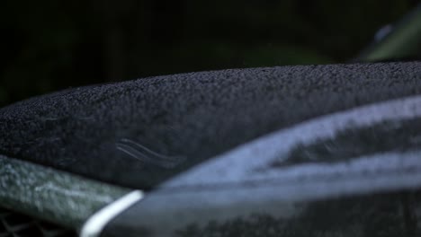 rain falls onto hood of black car and creates water droplets that run off, extreme close up, focus pull