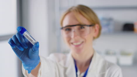 trabajadora de investigación de laboratorio sonriente con gafas de seguridad sosteniendo un tubo de ensayo etiquetado con vacuna