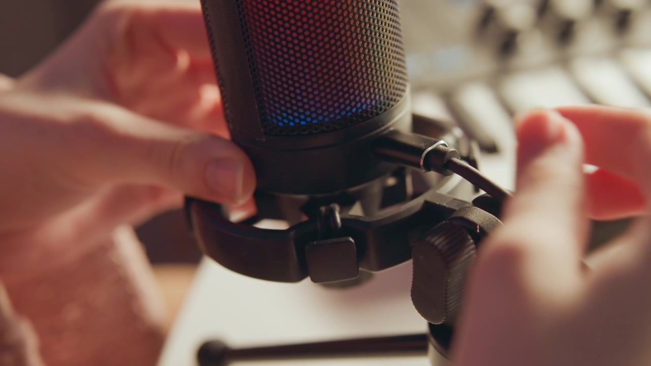 Close view of hands positioning condenser microphone inside shock mount in home studio, showing active finger movement, soft blur, glowing grille, warm light, and tidy recording workspace