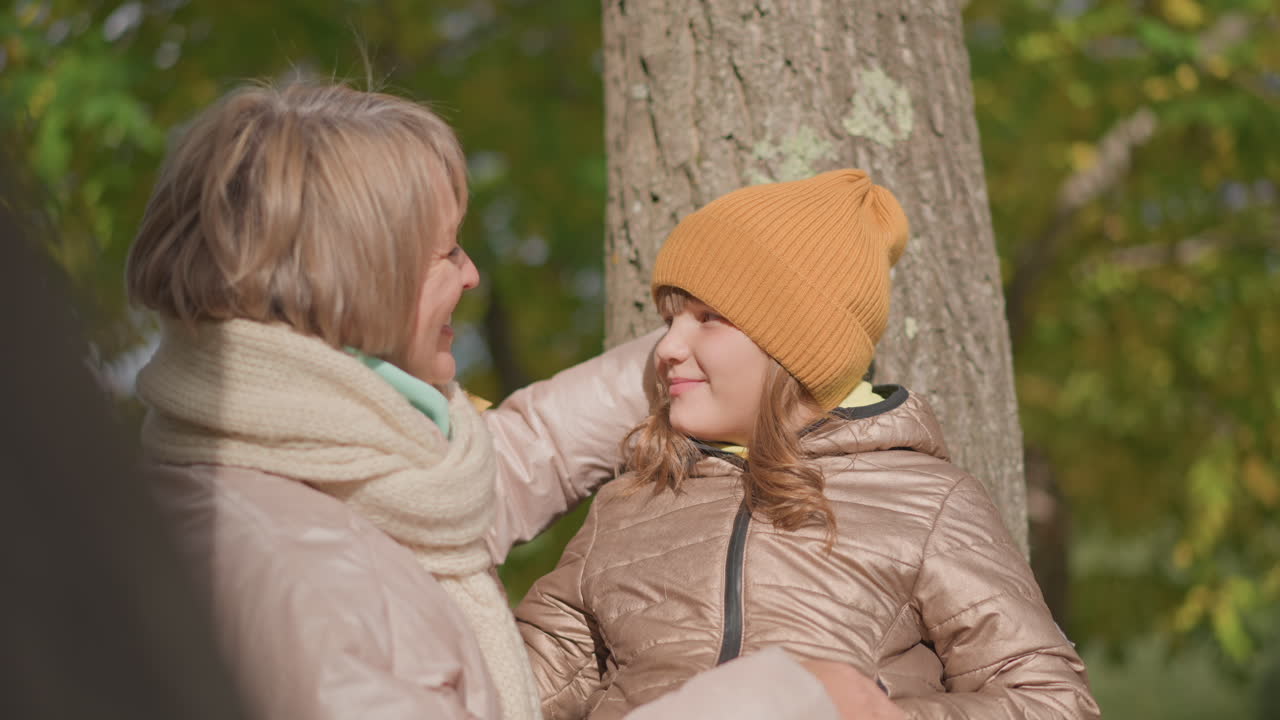 kid in mustard hat and shiny coat relaxes against tree trunk during sunny autumn day as loving mum offers bunch of yellow leaves and embraces her with warmth and gentle smile in peaceful outdoor park