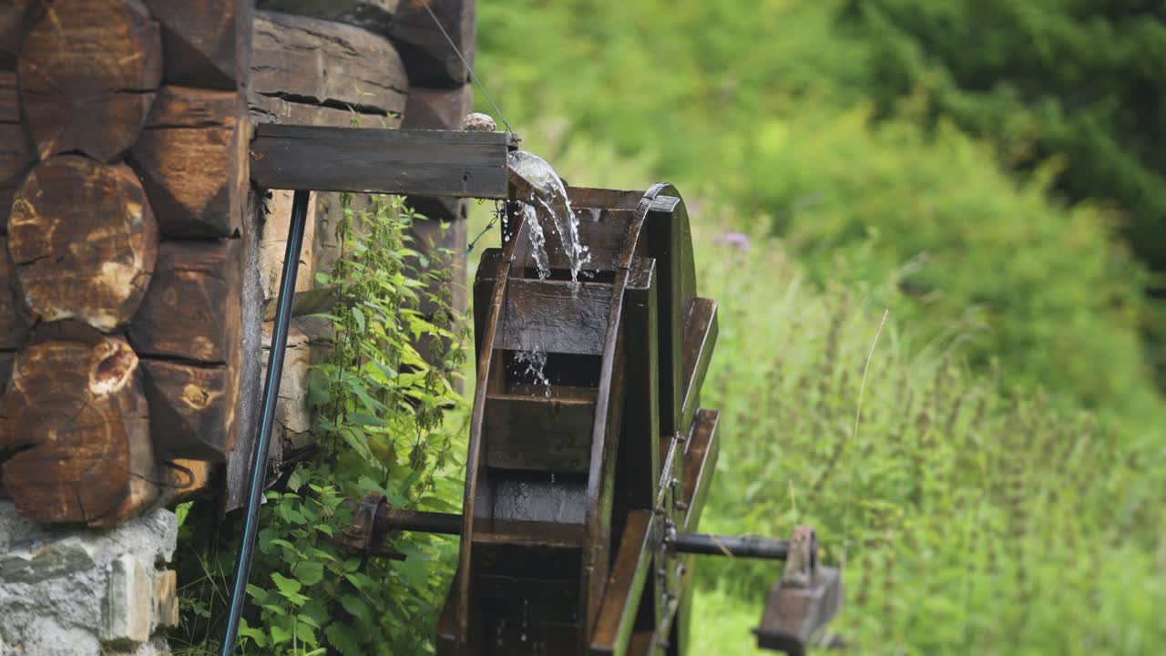 A wooden waterwheel turns as water flows onto it from a log chute, set against a lush green background.