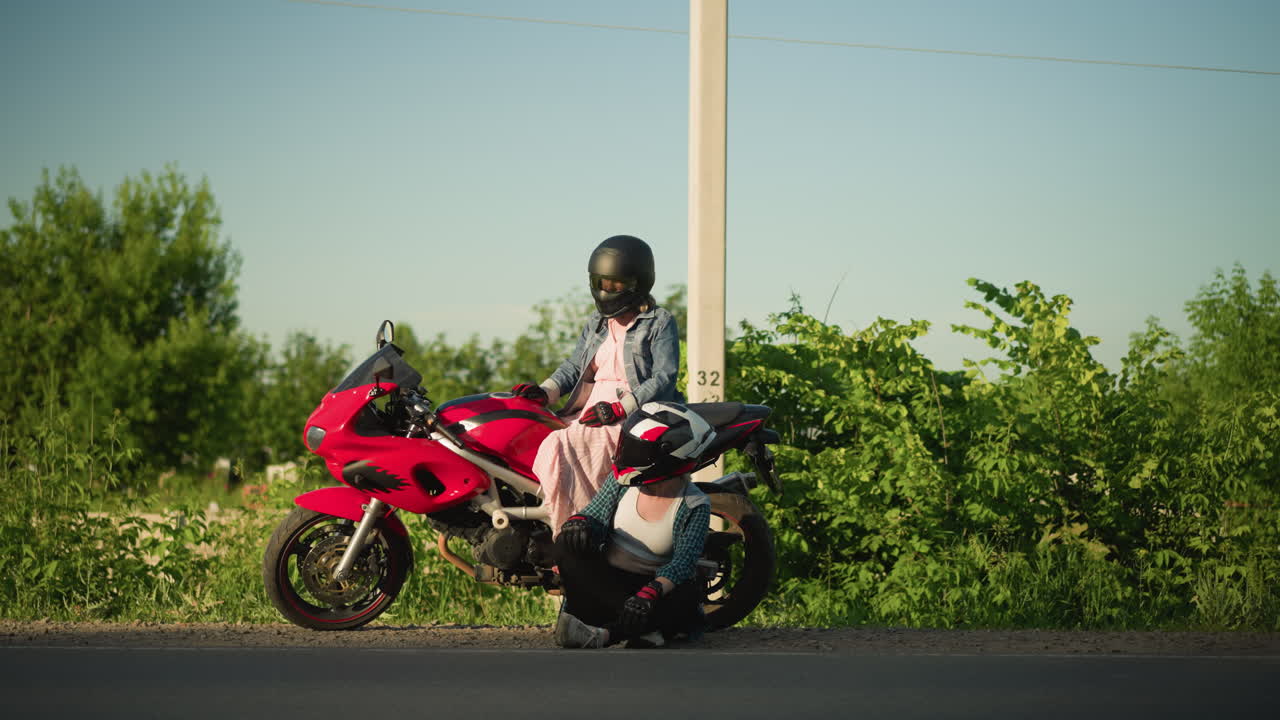 dos mujeres a la orilla de la carretera, una sentada en una motocicleta roja mirando a la izquierda y a la derecha mientras que la otra se sienta en el suelo, ambas mirando en la misma dirección