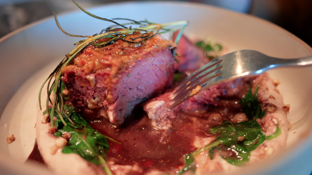 Close up of a woman's hand cutting into a gourmet meat dish served with sauce, greens, and rosemary garnish on a white plate