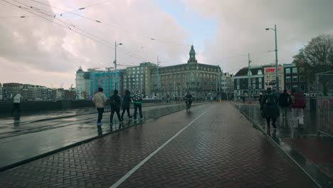 Senic view of Amsterdam's iconic brick road with pedestrians and cyclists passing by, surrounded by historic buildings, tram infrastructure, and cloudy sky after rainfall. Amsterdam, Netherlands