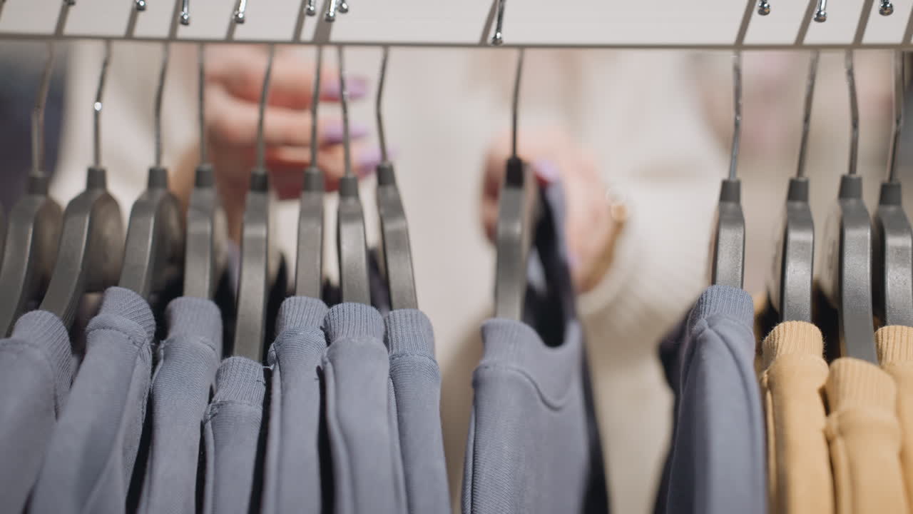 Close up view of clothes hanging on rack in bright store, blurred background shows female shopper hands gently sliding garments along hangers, fingernails painted pastel purple
