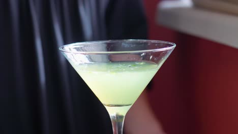 green cocktail with mint leaves is poured slowly into a martini glass closeup, alcoholic mixed drink is poured with cobbler shaker drips slowly from woman bartender’s hand
