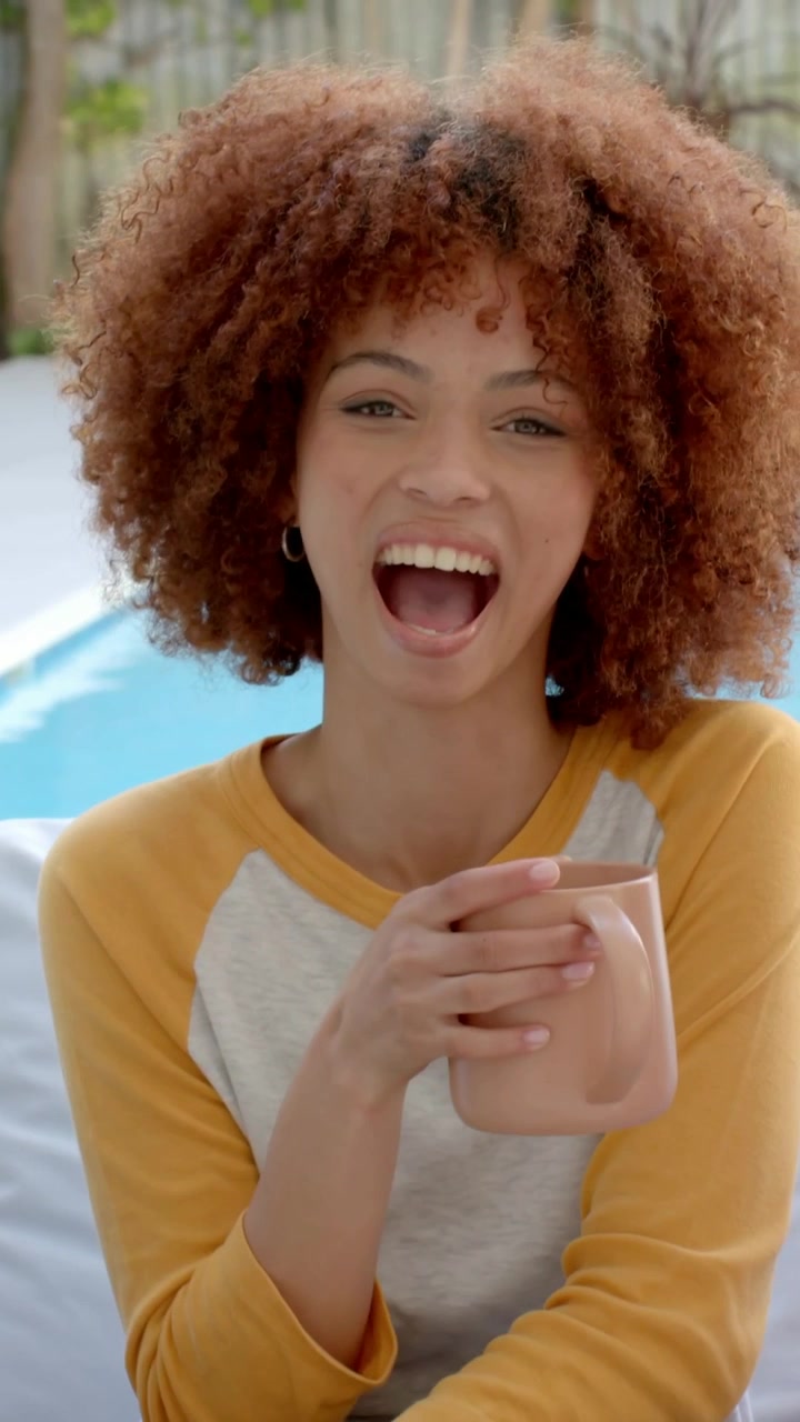 Video verticale: Donna sorridente in camicia color senape che tiene una tazza in piscina dopo un segnale fuori campo, ridendo.
