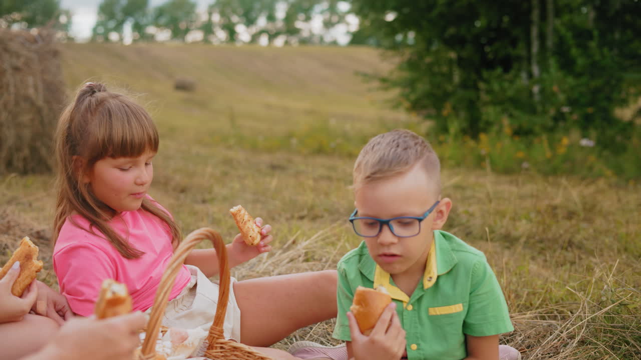 Famiglia serena seduta all'aperto che gusta spuntini mentre è circondata da una vasta campagna, i bambini mangiano felicemente dolci da un cestino da picnic mentre condividono un momento di relax nella natura.