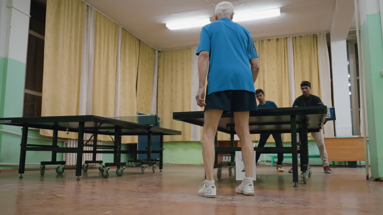 Elderly man in blue sportswear lunges forward to return table tennis ball during intense indoor rally as young opponents watch closely across net, dynamic action inside hall