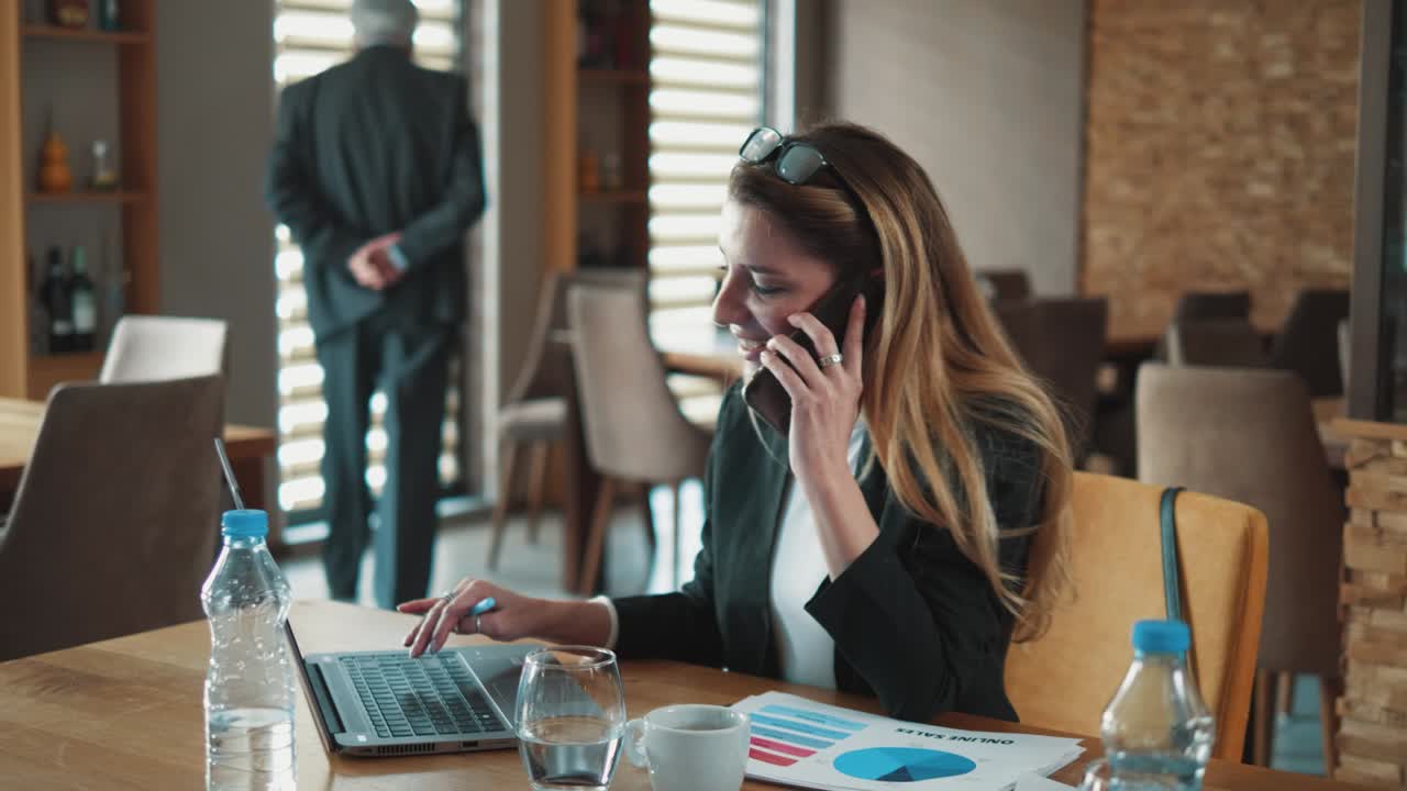 Ung forretningskvinde taler i telefon og arbejder på en bærbar computer, mens virksomhedens chef går i baggrunden