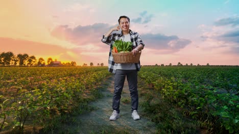 cuerpo lleno de granjero masculino asiático con canasta de verduras sonriendo cálidamente y haciendo hablar por teléfono gesto en el campo