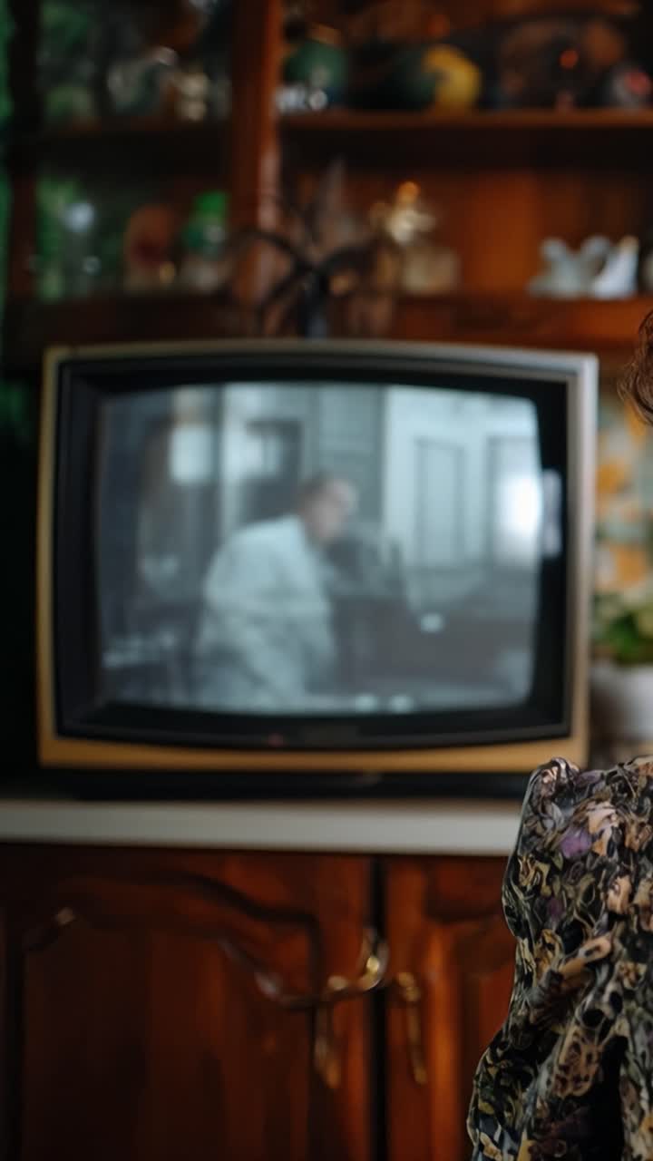 Žena sleduje starou televizi v útulném, teplém pokoji. Měkké světlo září na její vrásčitou tvář. Vintage televize ukazuje vybledlé rodinné scény. Teplé dřevěné skříňky rámují nostalgický okamžik. Její pohled skrývá tichou, něžnou zvědavost.