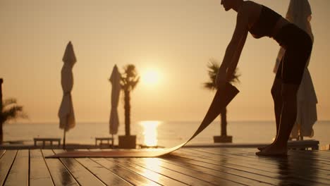 Primer-Plano-De-Una-Chica-Con-Un-Uniforme-Deportivo-Negro-De-Verano-Extendiendo-Un-Colchón-Para-Hacer-Yoga-Frente-A-Una-Puesta-De-Sol-Dorada-En-La-Playa.-Clases-De-Yoga-Y-Meditación-Por-La-Noche-En-Una-Playa-Soleada.