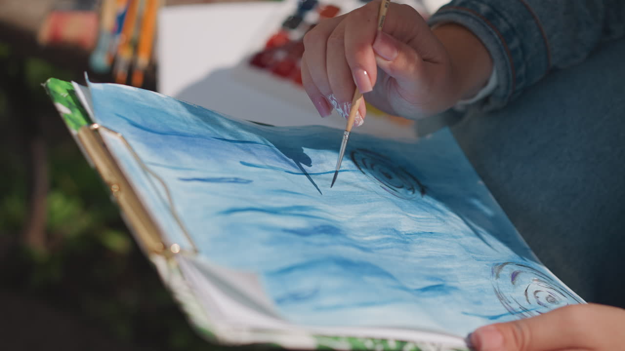 Blue watercolor seascape on clipboard, closeup of hands painting with fine brush, denim sleeve visible, natural sunlight, park background, wet washes and textured strokes conveying calm creative
