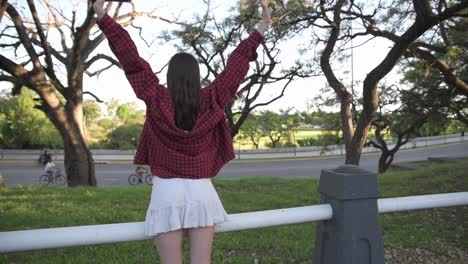 back view of beautiful woman raising her arms expressing freedom in park at sunset