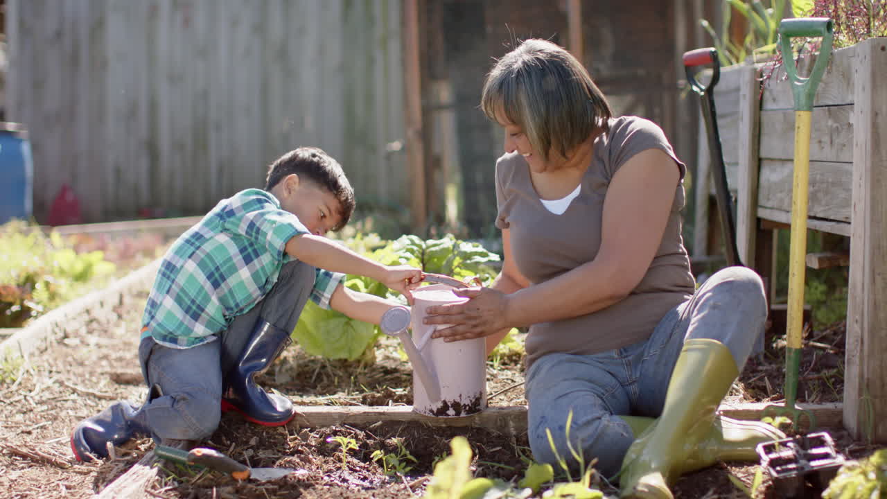 una abuela y un nieto bi-racial felices regando plantas en un jardín soleado, en cámara lenta