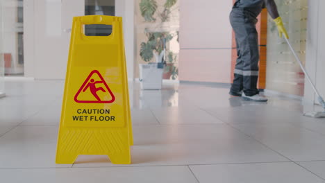 cleaning man cleaning the floor with mop inside an office building behind a wet floor warning sign