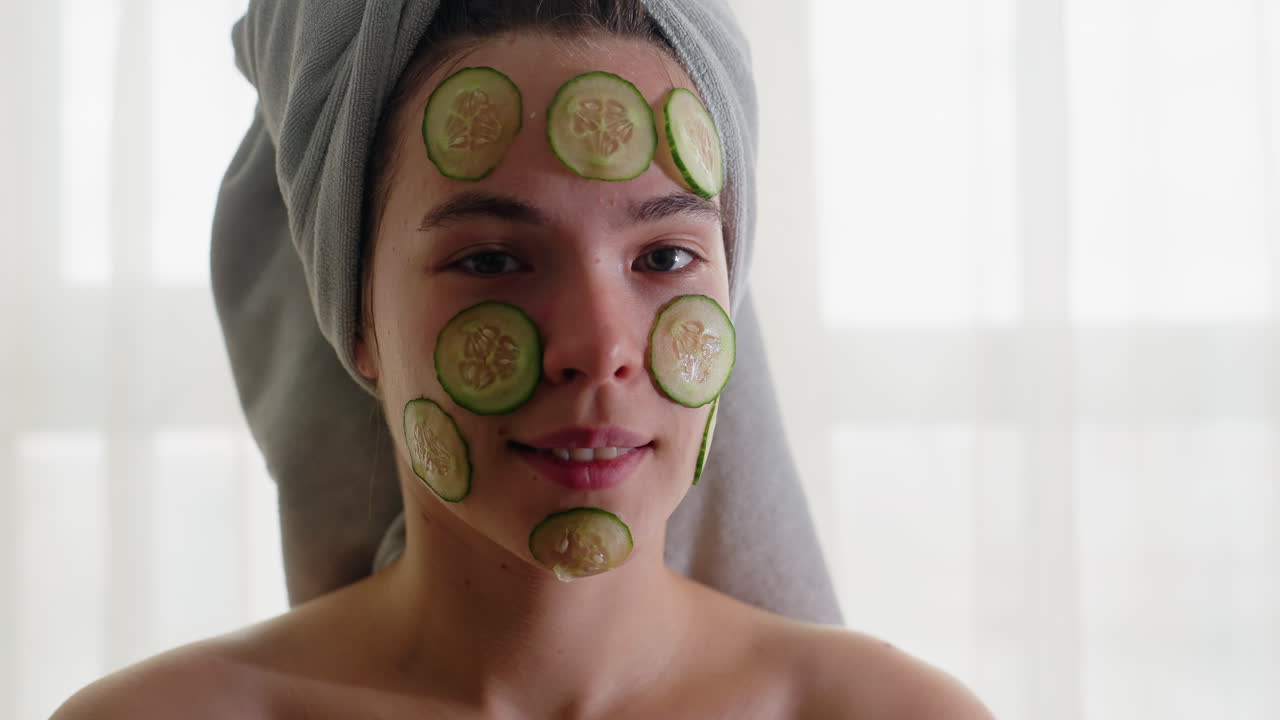 Portrait of woman with cucumber slices placed on face, towel wrapped around hair, blinking eyes during refreshing skincare treatment, enjoying rejuvenation, relaxation, and self-care routine