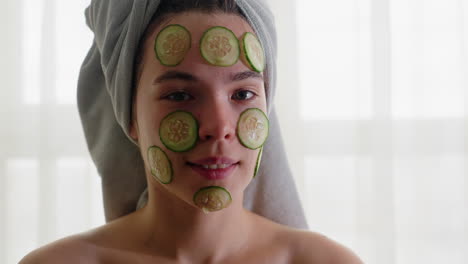 portrait of woman with cucumber slices placed on face, towel wrapped around hair, blinking eyes during refreshing skincare treatment, enjoying rejuvenation, relaxation, and self-care routine
