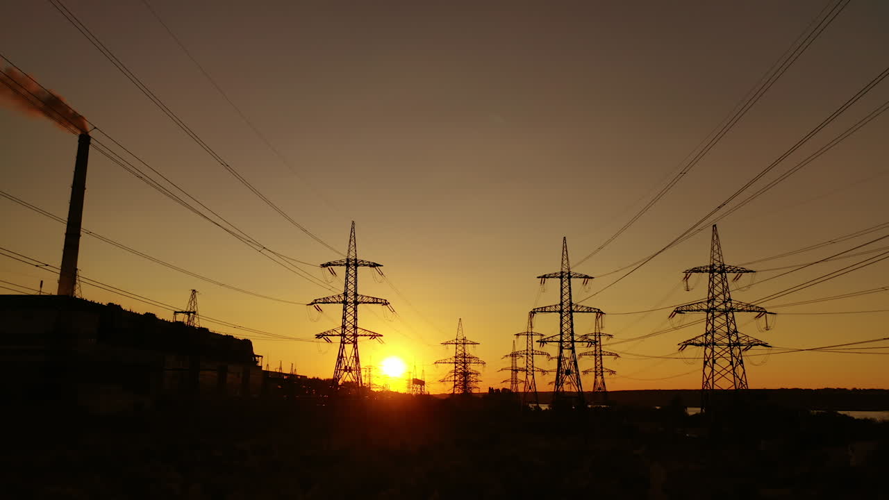 Transmission towers at dusk. Electric pylons near the smoking industrial pipe in the evening. Electric high voltage lines against beautiful orange sky at sunset. Energy efficiency conception.