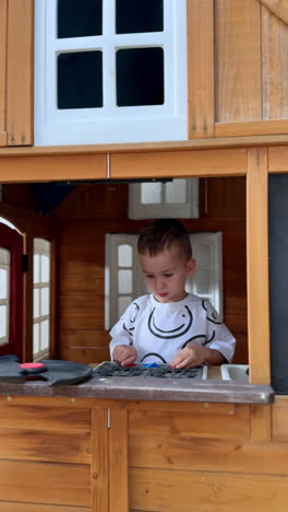 smiling happy kid stands at the window inside the wooden baby playhouse. baby boy plays with toy stove and waves hands joyfully. vertical video.