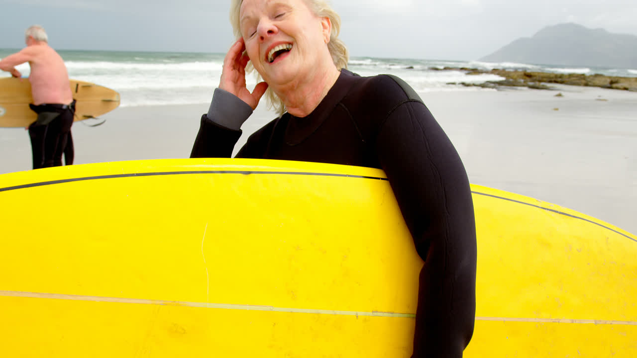 Front view of old caucasian woman holding surfboard at beach 4k