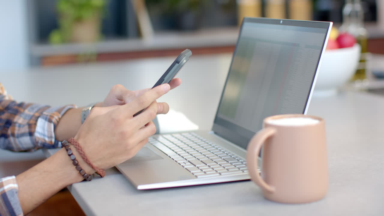 Using smartphone and laptop, teenage boy working from home with coffee mug nearby