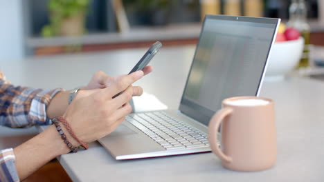 using smartphone and laptop, teenage boy working from home with coffee mug nearby