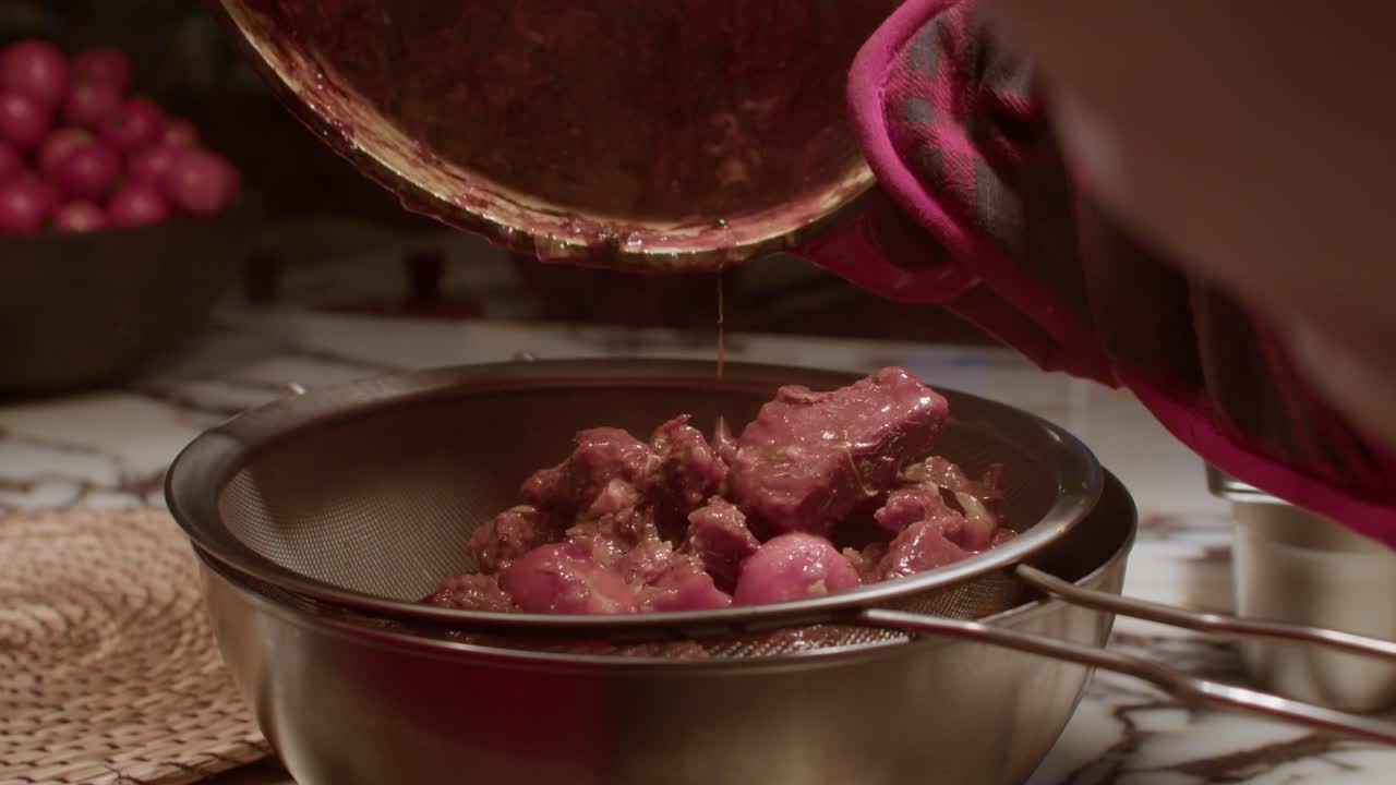 Gambar close-up detail seseorang yang menuangkan rebusan merah tua yang kaya dengan potongan daging dan sayuran besar dari panci ke dalam saringan logam