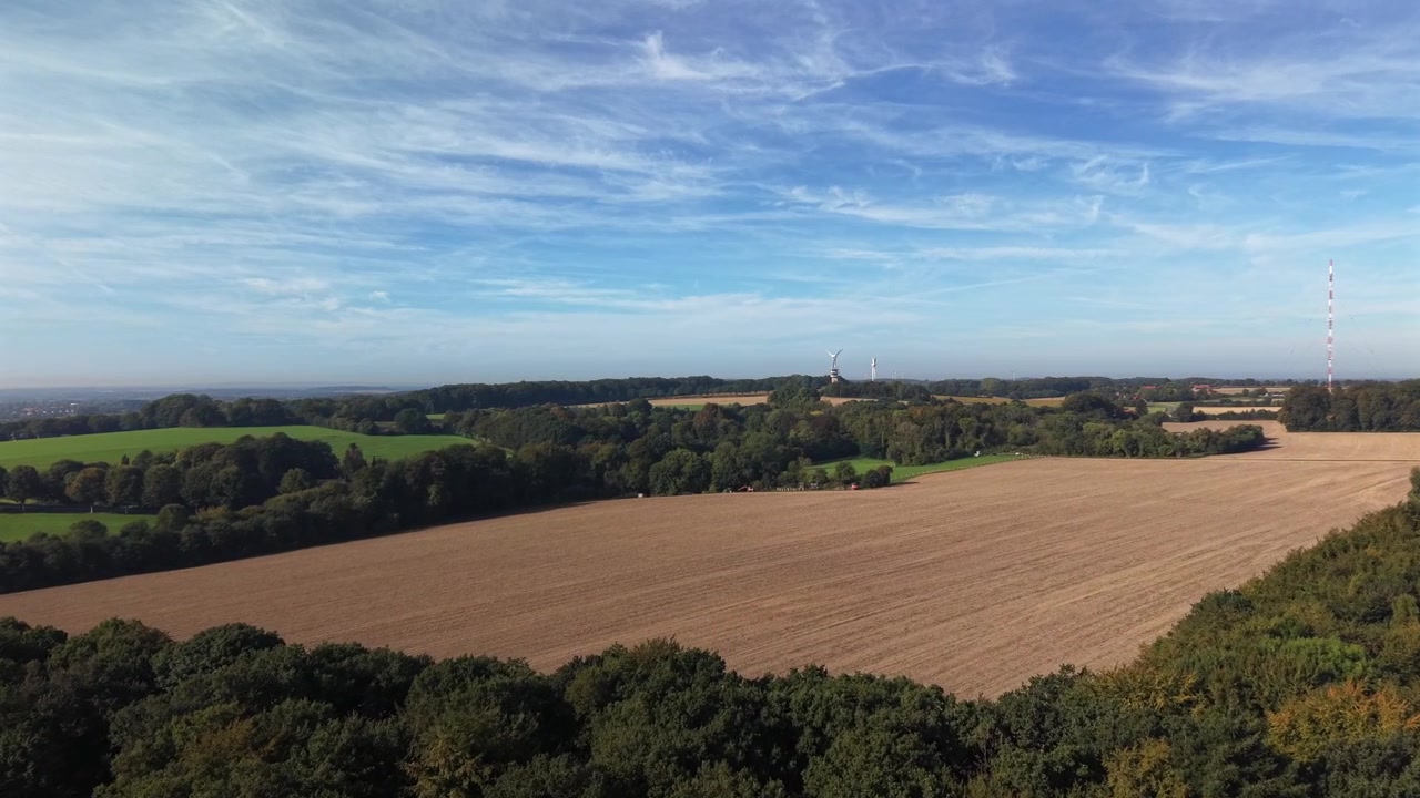 Calm autumn forest treetops above large harvested golden farm fields, red white radio tower, distant white wind turbines under blue sky with wispy clouds in rural America. Aerial drone wide shot