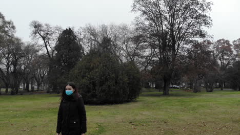 a young woman with a mask walks in a park green field on a cloudy day