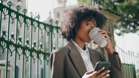 Successful-woman-scrolling-phone-holding-beverage-takeaway-outdoors-close-up.