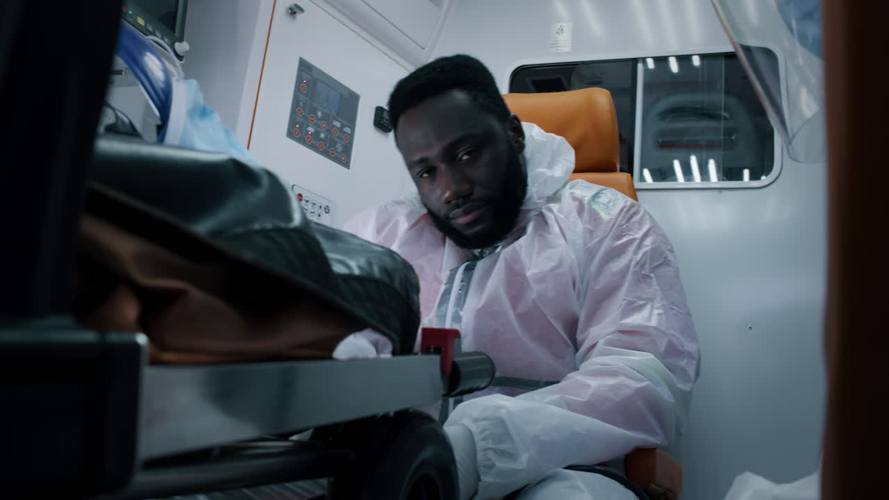 exhausted African American Paramedic Riding Ambulance Tired Black Doctor in Hazmat Suit Blinking Slowly and Wiping Sweat from Forehead Riding Ambulance to Hospital during Pandemic